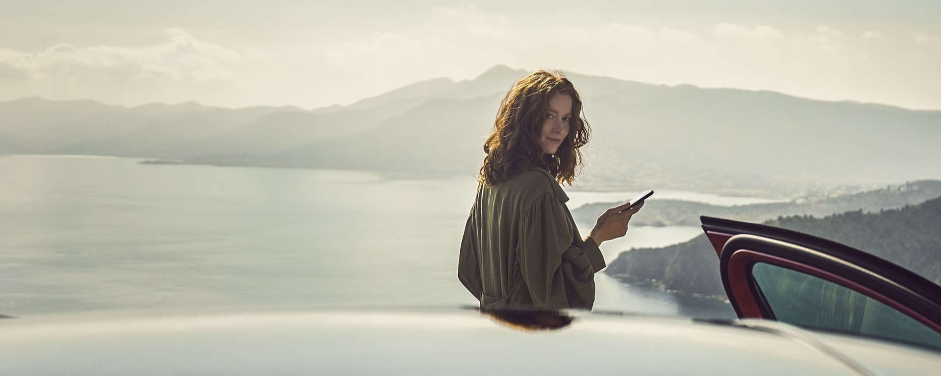 Дівчина зі смартфоном стоїть за автомобілем на фоні морської затоки